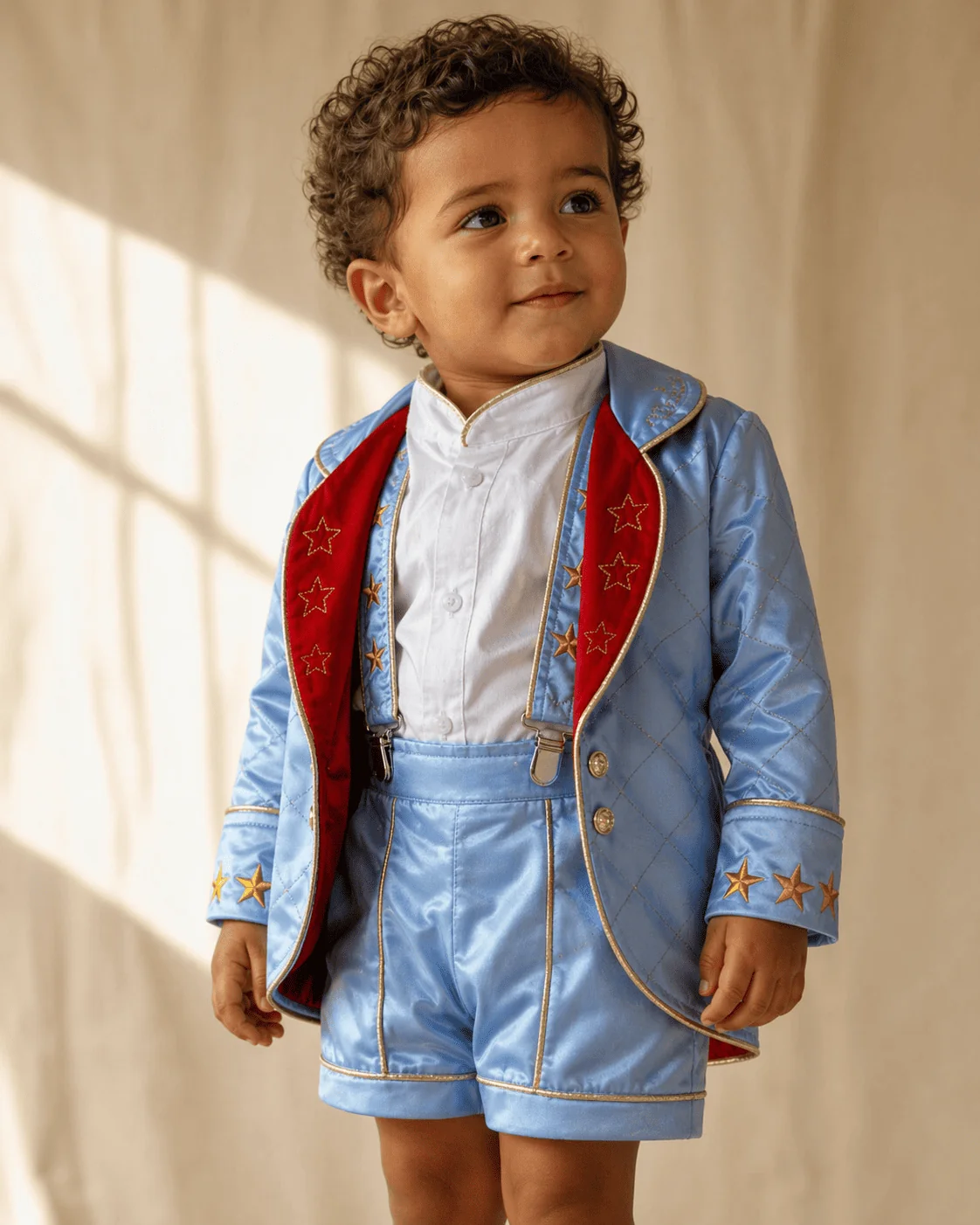 Menino brasileiro de 2 anos vestindo o Conjunto Príncipe Luxo completo — casaco de cetim azul claro com pesponto dourado em losangos, lapela de veludo vermelho com estrelas douradas bordadas, short coordenado e suspensório decorativo — retratado em pose três-quartos com luz natural quente, qualidade editorial de ateliê