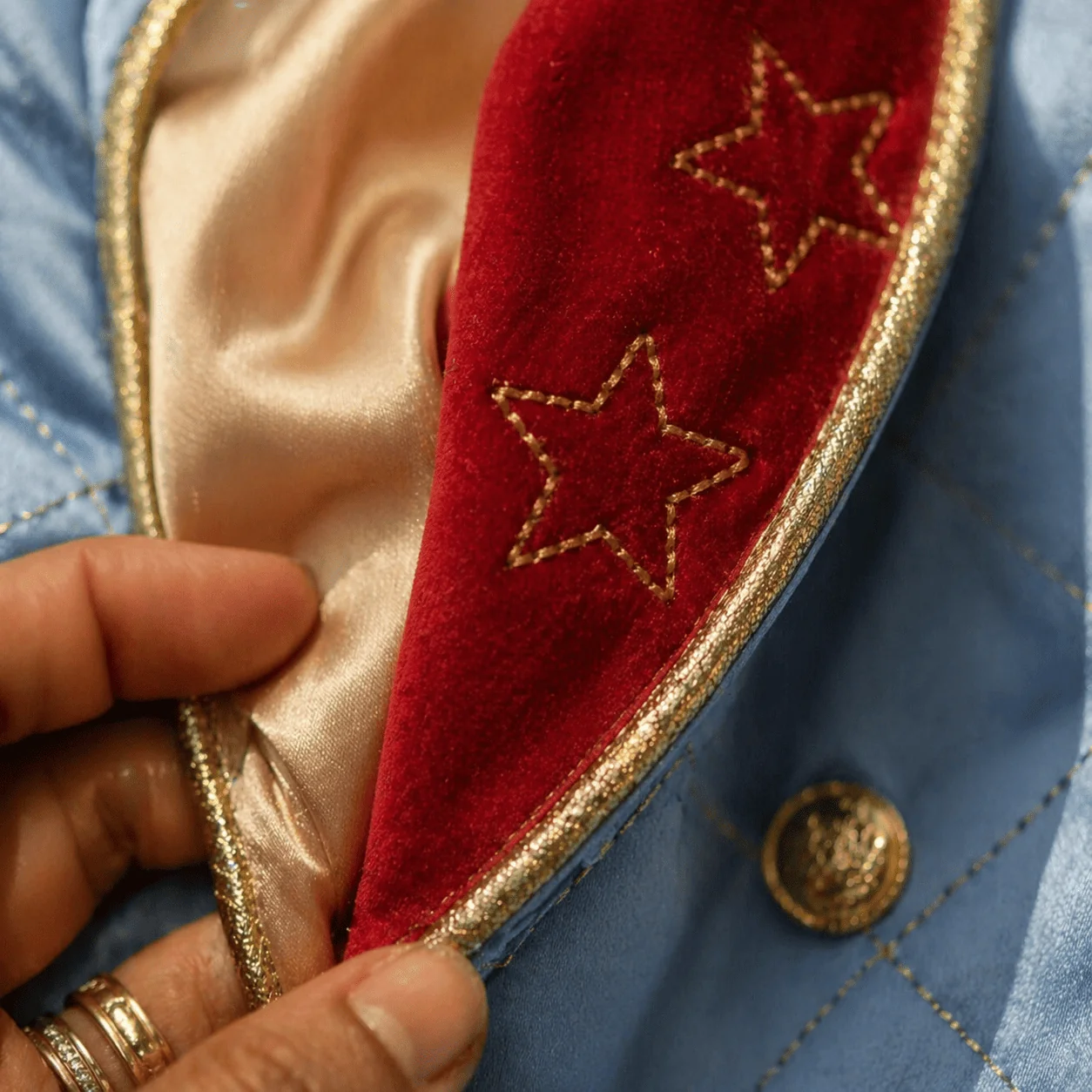 Macro extremo da lapela do casaco Príncipe Luxo aberta parcialmente revelando o forro em tricoline marfim por baixo — veludo vermelho com estrela dourada bordada encontra o cetim azul com pré-esponto quadriculado dourado, vivo dourado feito à mão na borda — mão feminina brasileira com aliança discreta segurando a lapela aberta
