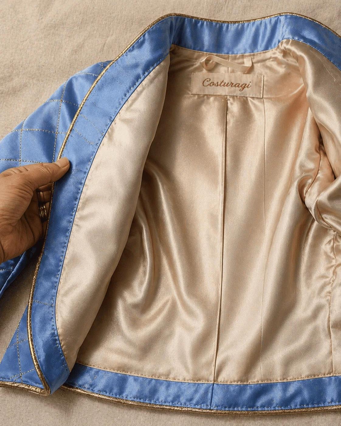 Casaco do Conjunto Príncipe Luxo aberto do avesso sobre superfície de linho cru marfim mostrando o forro em tricoline 100% algodão marfim em detalhe — costura invisível à mão na barra, costura interna limpa entre forro e entretela, alcinha de fita cetim no centro das costas com etiqueta Costuragi bordada em linha cobre — mão feminina virando uma lapela para revelar o acabamento museu-grade